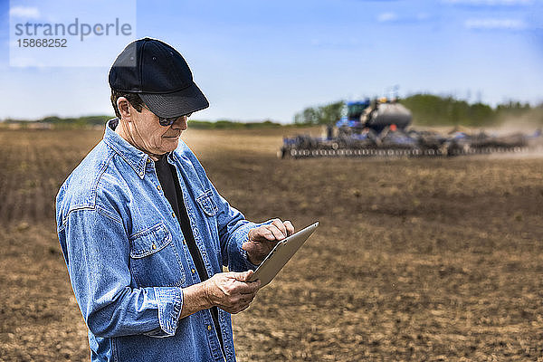 Landwirt  der ein Tablet benutzt  während er auf einem landwirtschaftlichen Feld steht und ein Traktor und Geräte das Feld besäen; Alberta  Kanada