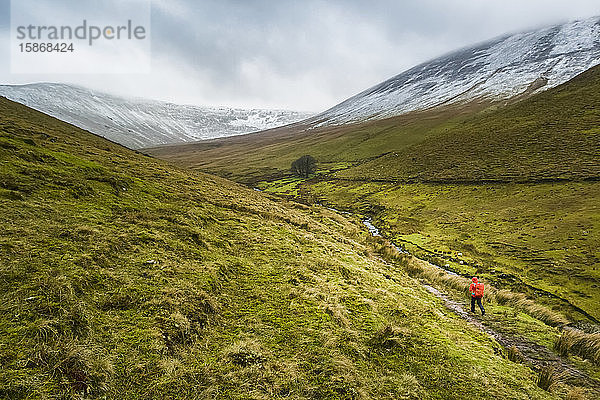 Einsame Wanderin in rotem Regenmantel mit rotem Rucksack  die an einem bewölkten Wintertag einen Talweg entlanggeht  der zu schneebedeckten Bergen führt  Galty Mountains; County Tipperary  Irland