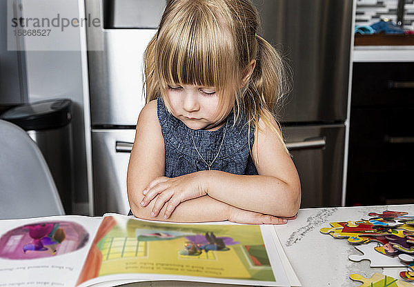 Ein süßes kleines Mädchen  das ein Bilderbuch liest und ein Puzzle auf einem Tisch in der Küche macht: Edmonton  Alberta  Kanada