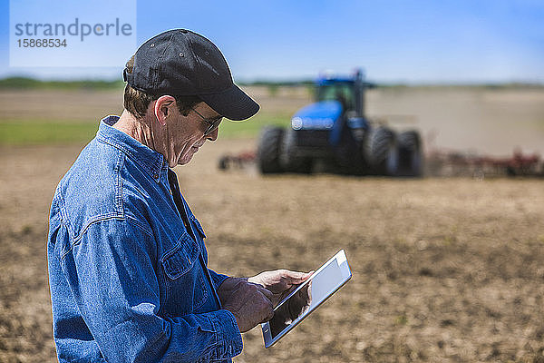 Landwirt  der ein Tablet benutzt  während er auf einem landwirtschaftlichen Feld steht und ein Traktor und Geräte das Feld besäen; Alberta  Kanada