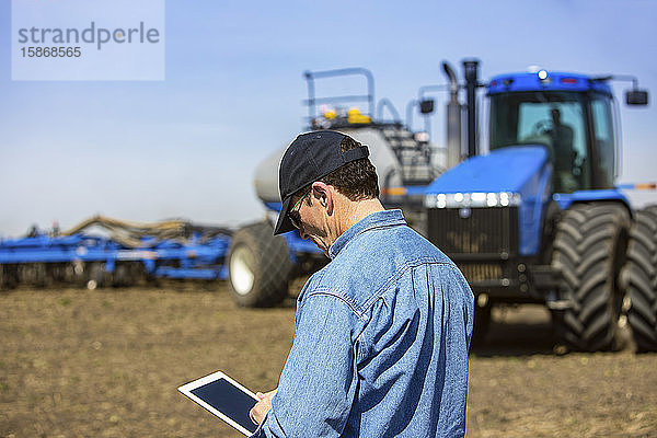 Landwirt  der ein Tablet benutzt  während er auf einem landwirtschaftlichen Feld steht und ein Traktor und Geräte das Feld besäen; Alberta  Kanada