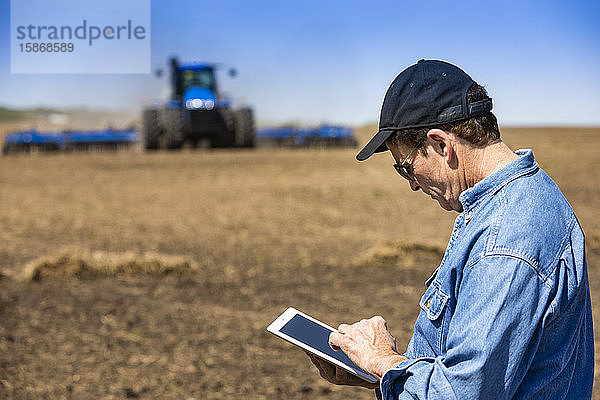 Landwirt  der ein Tablet benutzt  während er auf einem landwirtschaftlichen Feld steht und ein Traktor und Geräte das Feld besäen; Alberta  Kanada