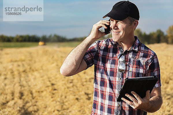 Ein Landwirt telefoniert und nutzt sein Tablet  um bei der Weizenernte zu helfen  während im Hintergrund ein Mähdrescher arbeitet: Alcomdale  Alberta  Kanada