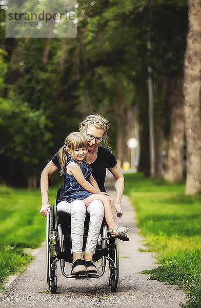Eine querschnittsgelähmte Mutter hält ihr kleines Mädchen auf dem Schoß  während sie es an einem warmen Sommernachmittag in ihrem Rollstuhl die Straße hinunterfährt: Edmonton  Alberta  Kanada.