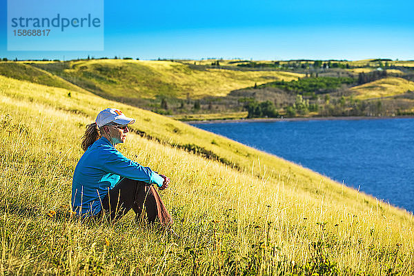 Frau sitzt auf einem grasbewachsenen Hügel mit Blick auf einen See und sanfte Hügel im Hintergrund mit blauem Himmel  Waterton Lakes National Park; Waterton  Alberta  Kanada