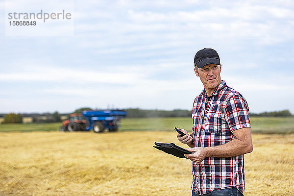 Ein Landwirt nutzt sein Tablet  um die Weizenernte zu verwalten  während im Hintergrund ein Getreidewagen arbeitet; Alcomdale  Alberta  Kanada