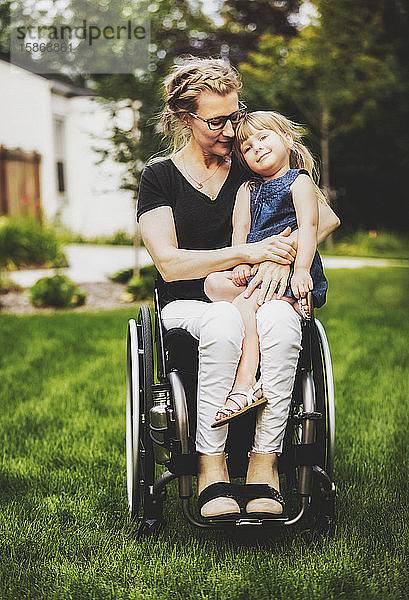 Eine querschnittsgelähmte Mutter hält ihr kleines Mädchen auf dem Schoß  während sie an einem warmen Sommernachmittag in ihrem Vorgarten im Rollstuhl sitzt: Edmonton  Alberta  Kanada.