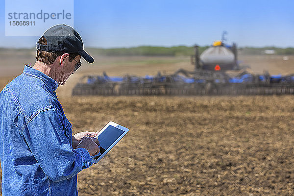 Landwirt  der ein Tablet benutzt  während er auf einem landwirtschaftlichen Feld steht und ein Traktor und Geräte das Feld besäen; Alberta  Kanada