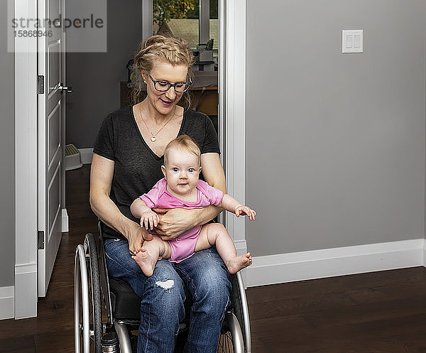 Eine querschnittsgelähmte Mutter  die ihr Baby auf dem Schoß hält  während sie sich im Rollstuhl durch ihr Haus bewegt:: Edmonton  Alberta  Kanada