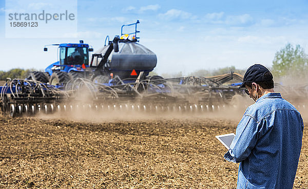 Landwirt  der ein Tablet benutzt  während er auf einem landwirtschaftlichen Feld steht und ein Traktor und Geräte das Feld besäen; Alberta  Kanada