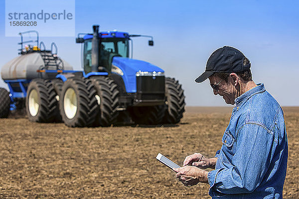 Landwirt  der ein Tablet benutzt  während er auf einem landwirtschaftlichen Feld steht und ein Traktor und Geräte das Feld besäen; Alberta  Kanada