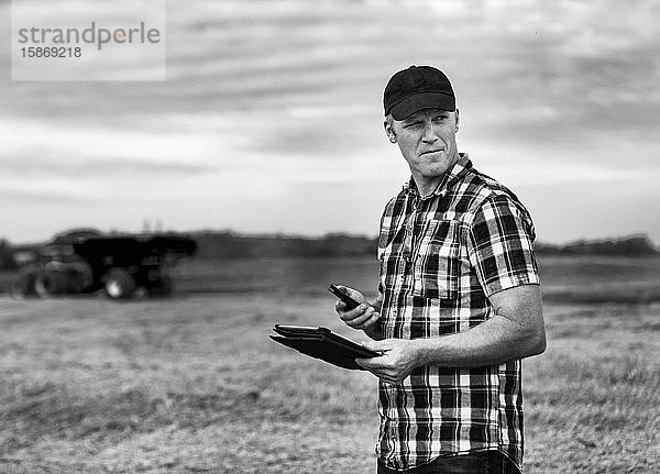 Ein Landwirt nutzt sein Tablet  um die Weizenernte zu verwalten  während im Hintergrund ein Getreidewagen arbeitet: Alcomdale  Alberta  Kanada