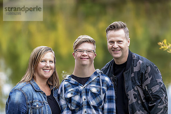 Ein junger Mann mit Down-Syndrom posiert für ein Familienporträt mit seinem Vater und seiner Mutter  während sie an einem warmen Herbstabend in einem Stadtpark die Gesellschaft des jeweils anderen genießen: Edmonton  Alberta  Kanada