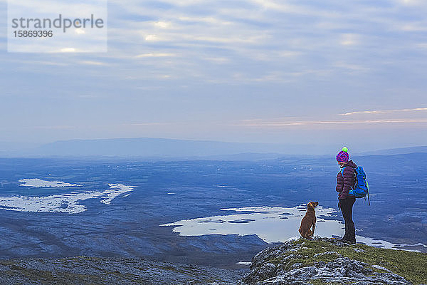 Einsame Wanderin mit Strickmütze und Hund  die sich an einem bewölkten Abend im Winter auf einer Klippe mit Blick auf Seen in der Ferne ansehen  Burren National Park; County Clare  Irland