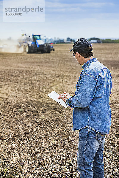 Landwirt  der ein Tablet benutzt  während er auf einem landwirtschaftlichen Feld steht und ein Traktor und Geräte das Feld besäen; Alberta  Kanada