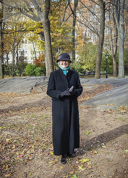 Ältere Frau steht für ein Porträt im Central Park im Herbst  hält ein Smartphone und trägt einen langen Mantel; New York City  New York  Vereinigte Staaten von Amerika