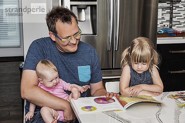 Ein Vater setzt sich mit seiner Tochter in die Küche  um ein Buch zu lesen und ein Puzzle zu bauen  während er sein Baby auf dem Schoß hält: Edmonton  Alberta  Kanada