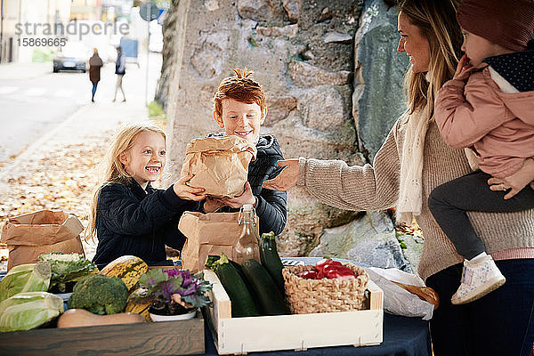 Lächelnde männliche und weibliche Geschwister kaufen Gemüse von weiblichen Marktverkäufern