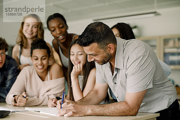 Lächelnder Tutor unterrichtet jugendliche Schüler  während er sich im Klassenzimmer über den Tisch lehnt