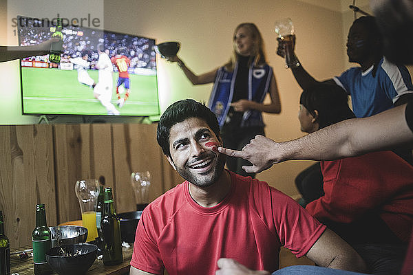Geschnittene Hand eines Mannes trägt während eines Fussballspiels Gesichtsfarbe auf die Wange eines Freundes auf