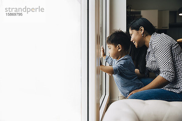 Seitenansicht von Mutter und Sohn beim Blick durchs Fenster  während sie zu Hause sitzen