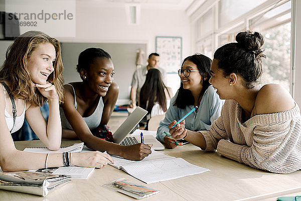 Lächelnde weibliche Teenager diskutieren  während sie im Klassenzimmer am Tisch sitzen