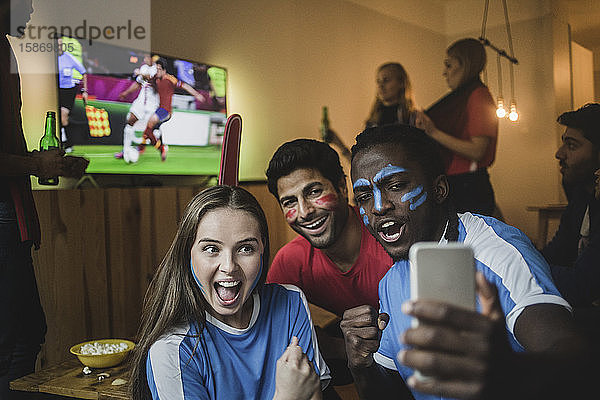 Fans nehmen Selfie mit dem Handy  während Freunde ein Fußballspiel im Fernsehen anschauen