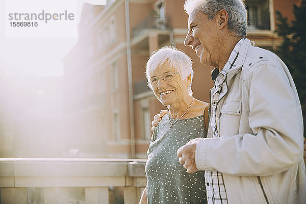 Lächelndes älteres Paar mit umarmtem Arm an einem sonnigen Tag in der Stadt spazieren
