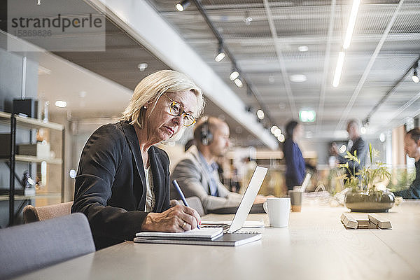 Senior-Unternehmerin schreibt Tagebuch  während sie im Büro am Tisch arbeitet