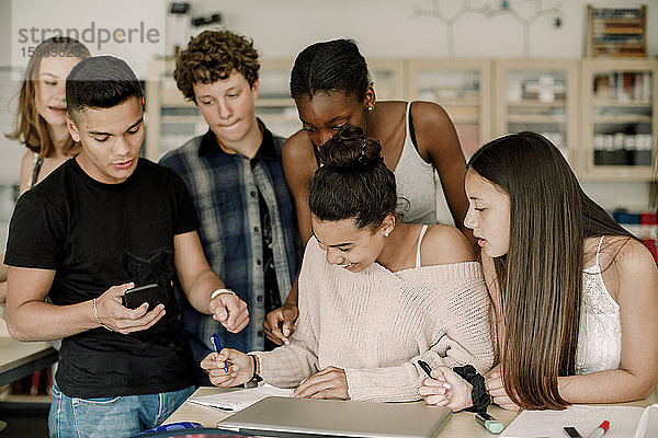 Lächelnder Teenager lernt  während Freunde im Klassenzimmer am Tisch stehen
