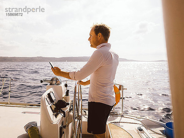 Seitenansicht eines Mannes  der sein Mobiltelefon in der Hand hält  während er bei strahlendem Sonnenschein auf einem Katamaran auf dem Meer gegen den Himmel segelt