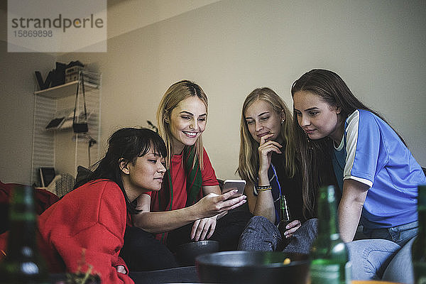 Lächelnde Freundinnen  die ein Mobiltelefon benutzen  während sie zu Hause sitzen