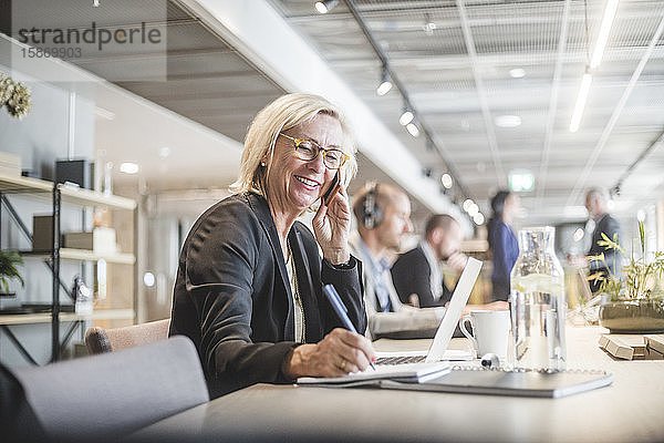 Lächelnde Geschäftsfrau  die während der Arbeit am Büroschreibtisch mit dem Handy telefoniert und in ein Tagebuch schreibt