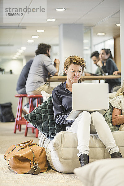 Unternehmerin arbeitet am Laptop  während ihre Mitarbeiter im Hintergrund sitzen