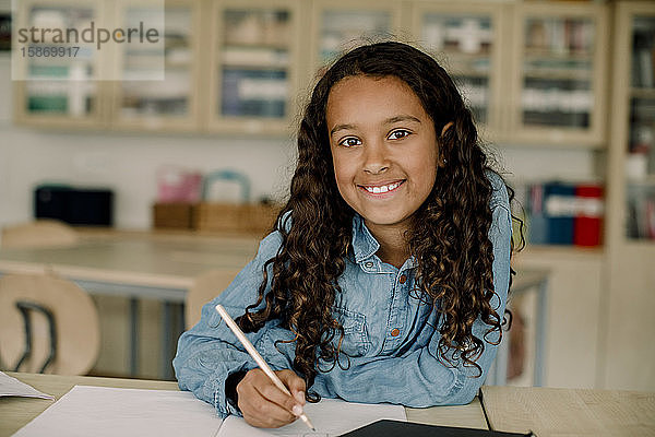 Porträt eines lächelnden Schülers  der in einem Buch schreibt  während er im Klassenzimmer am Tisch sitzt