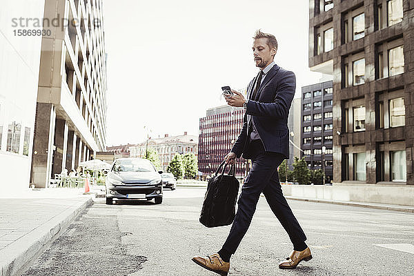 Männlicher Unternehmer  der beim Überqueren der Straße auf das Telefon schaut