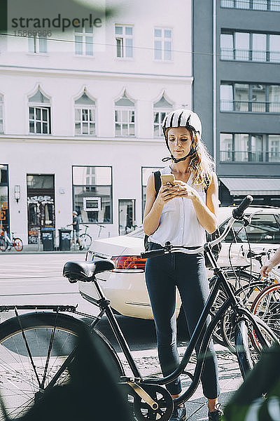 Geschäftsfrau mit Helm  die mit ihrem Mobiltelefon auf der Straße in der Stadt steht