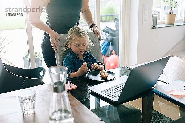 Mädchen isst Obst  während die Mutter zu Hause hinter einem Stuhl steht