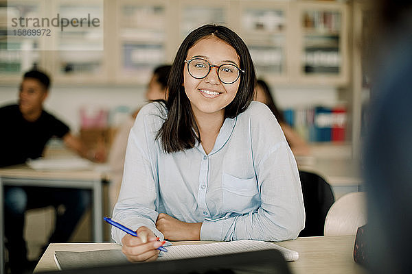 Porträt einer lächelnden Teenagerin mit Brille beim Lernen am Tisch im Klassenzimmer