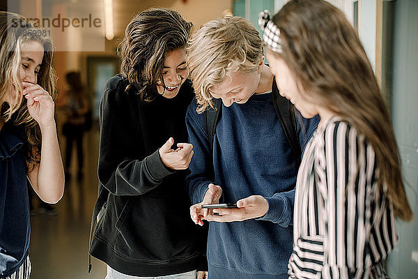 Lächelnde männliche Schüler  die ein Smartphone benutzen  während sie im Schulkorridor bei Freundinnen stehen