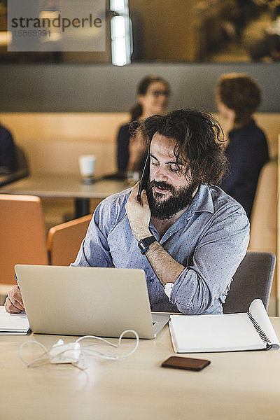 Männlicher Unternehmer mit Laptop  der mit seinem Handy telefoniert  während er im Büro am Tisch sitzt