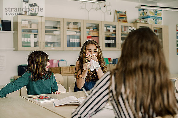 Lächelnde Studentin hält Papierbasteleien in der Hand  während sie im Klassenzimmer bei einem Freund sitzt