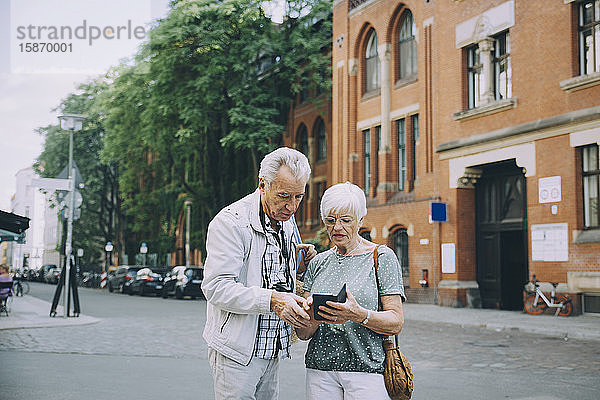 Ältere Frau hält Smartphone in der Hand und diskutiert mit Partner während einer Erkundungstour in der Stadt