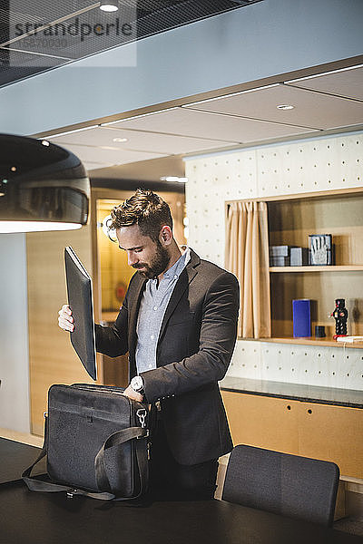 Junger Geschäftsmann hält Laptop in Tasche  während er im Büro am Tisch steht
