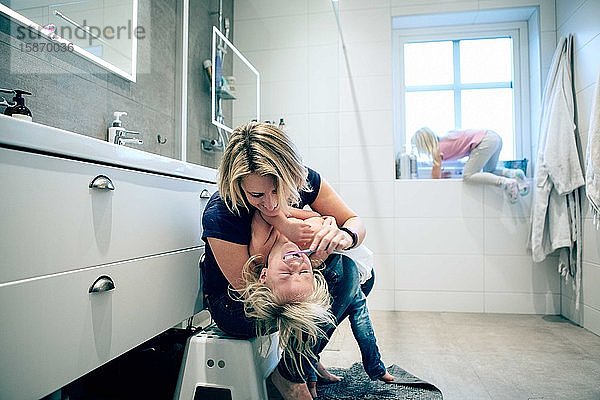 Lächelnde Mutter putzt der Tochter die Zähne  während das Mädchen im Badezimmer auf das Fenster klettert