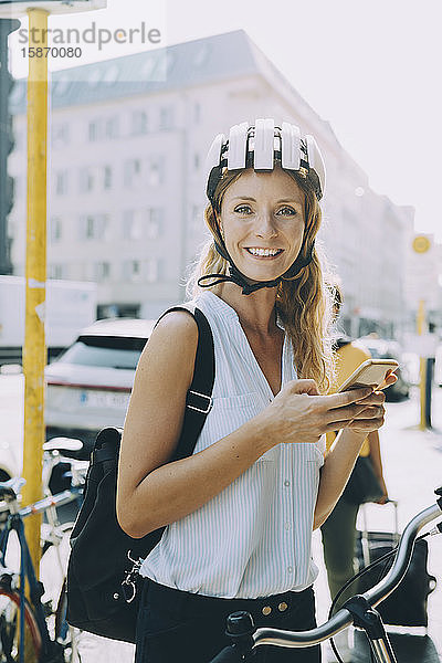 Porträt einer lächelnden Geschäftsfrau  die ein Mobiltelefon benutzt  während sie in der Stadt steht