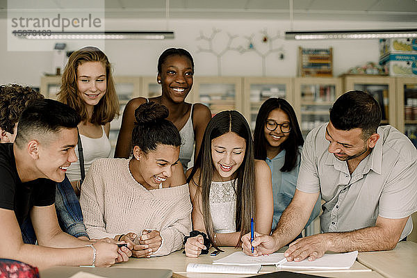 Lächelnder Tutor unterrichtet männliche und weibliche Studenten  während er sich im Klassenzimmer über den Tisch lehnt