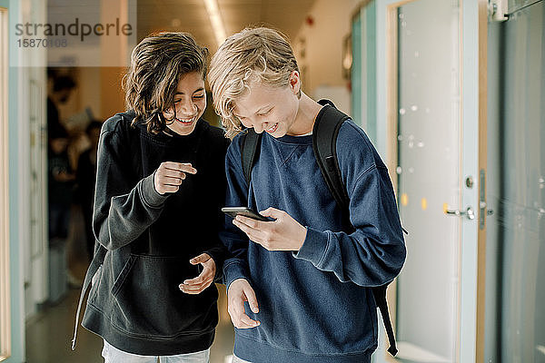 Lächelnde männliche Schüler mit Smartphone auf dem Schulkorridor