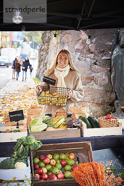 Porträt einer lächelnden Frau  die am Marktstand einen Korb mit frischen Kartoffeln hält