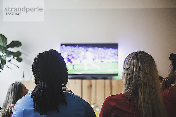 Rückansicht von Freunden  die sich ein Fußballspiel ansehen  während sie im Wohnzimmer sitzen